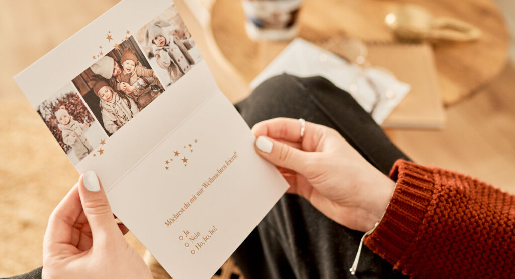 Eine Frau hält eine aufgeklappte Karte in der Hand. Im oberen Teil der Karte sind Fotos zu sehen, unten eine Einladung zu Weihnachten.