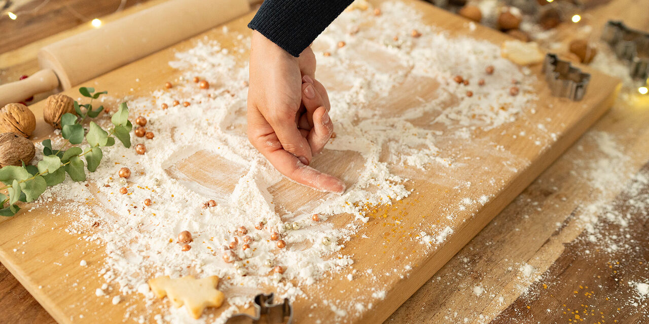 Auf dem Foto ist ein großes, mit Mehl bestreutes Holzbrett zu sehen. Der Rand des Brettes wurde weihnachtlich mit Ausstechformen, Keksen und Nüssen dekoriert. Zudem ist eine Hand zu erkennen, die mit dem Zeigefinger die Form eines Engels in das Mehl malt.