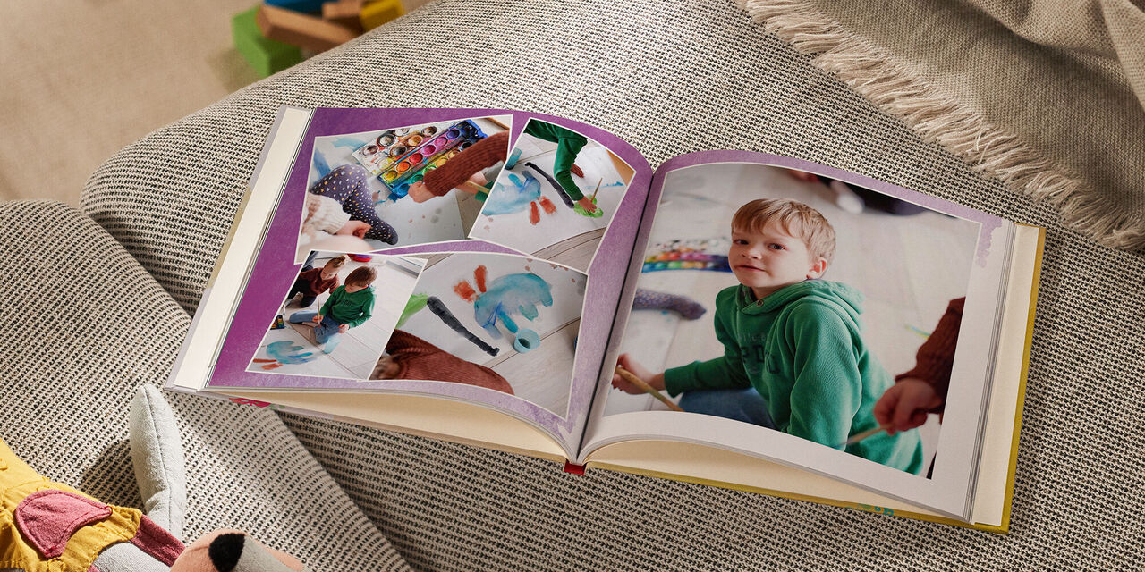 Auf einem Sofa liegt ein geöffnetes CEWE FOTOBUCH neben einem Kuscheltier und einer Decke. Auf der linken Seite sind vier Fotos zu erkennen, die sich leicht überlappen. Alle zeigen ein Kind, das mit den Farben aus einem Tuschkasten ein Bild malt. Die rechte Seite zeigt ein großes Foto von einem Jungen, der in die Kamera schaut. Die Doppelseite ist mit einem violetten Aquarellmuster hinterlegt.