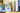 Auf einem fliederfarbenen Regal mit gelbem Hintergrund stehen verschiedene CEWE FOTOBUCH Exemplare von Reisen. Der Titel eines Buches zeigt das Foto eines Paares. Davor steht eine Bilderbox, die das Motiv einer Frau vor einem blauen Himmel zeigt.